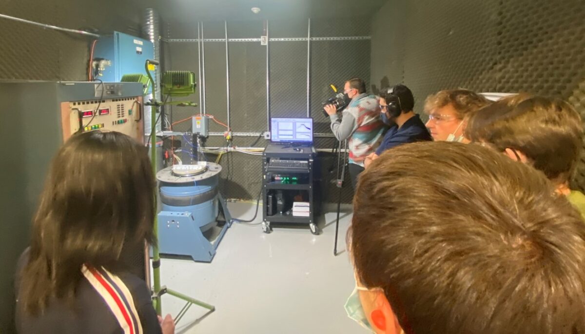 students and cameramen watch black satellite vibrate on blue G-force simulator connected to black computer