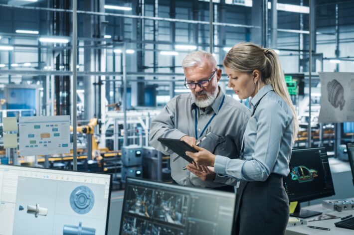 Two automotive engineers stand discussing fatigue testing results in a glass-walled room with several computer monitors in view