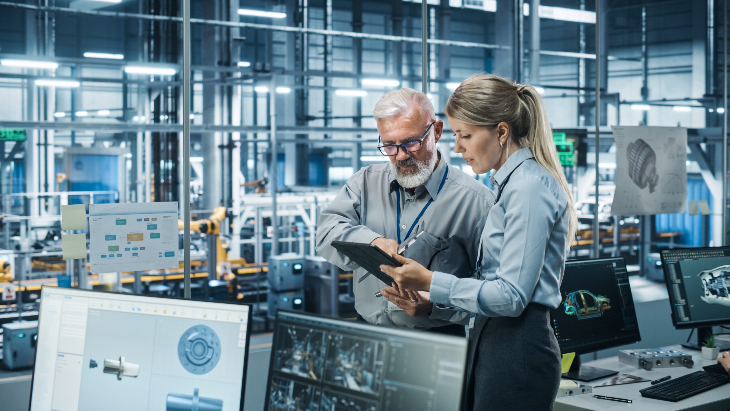 Two automotive engineers stand discussing fatigue testing results in a glass-walled room with several computer monitors in view