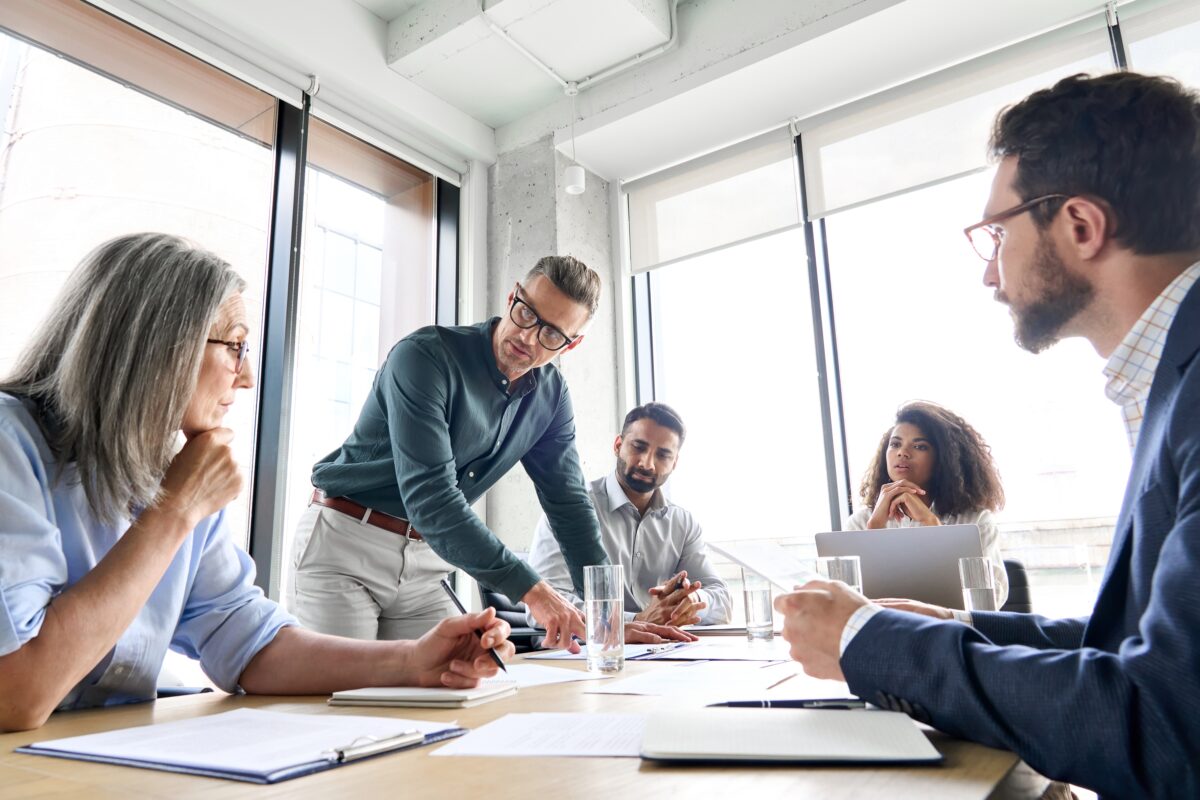A team of product engineers meets in a sunny conference room with papers on the wood-topped table