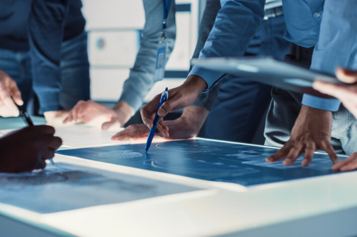 A group of engineers’ arms and hands are visible as they gather around plans for fatigue testing procedures. In the center, a hand holding a pencil points to plans on the worktable.