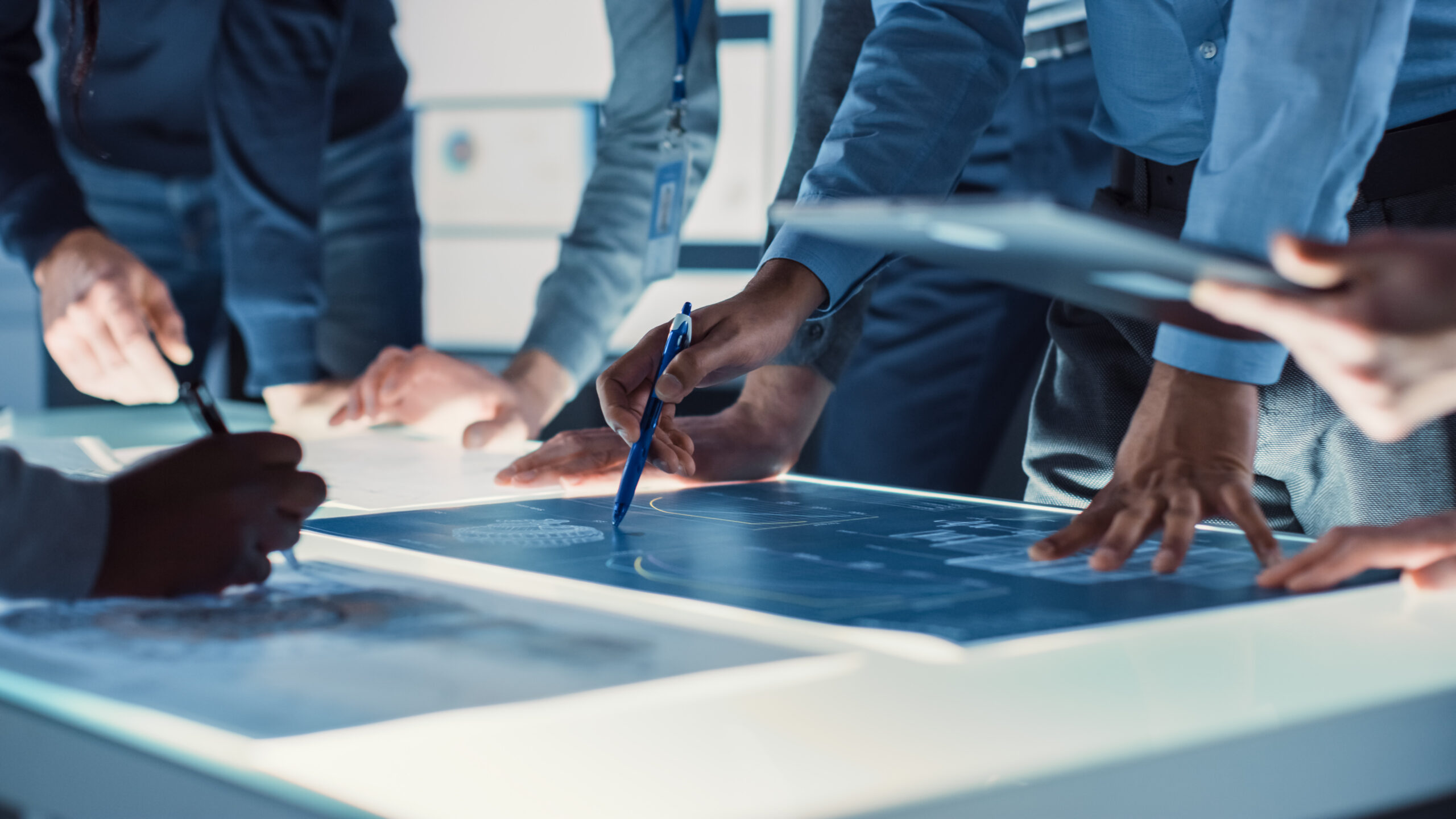 A group of engineers’ arms and hands are visible as they gather around plans for fatigue testing procedures. In the center, a hand holding a pencil points to plans on the worktable.
