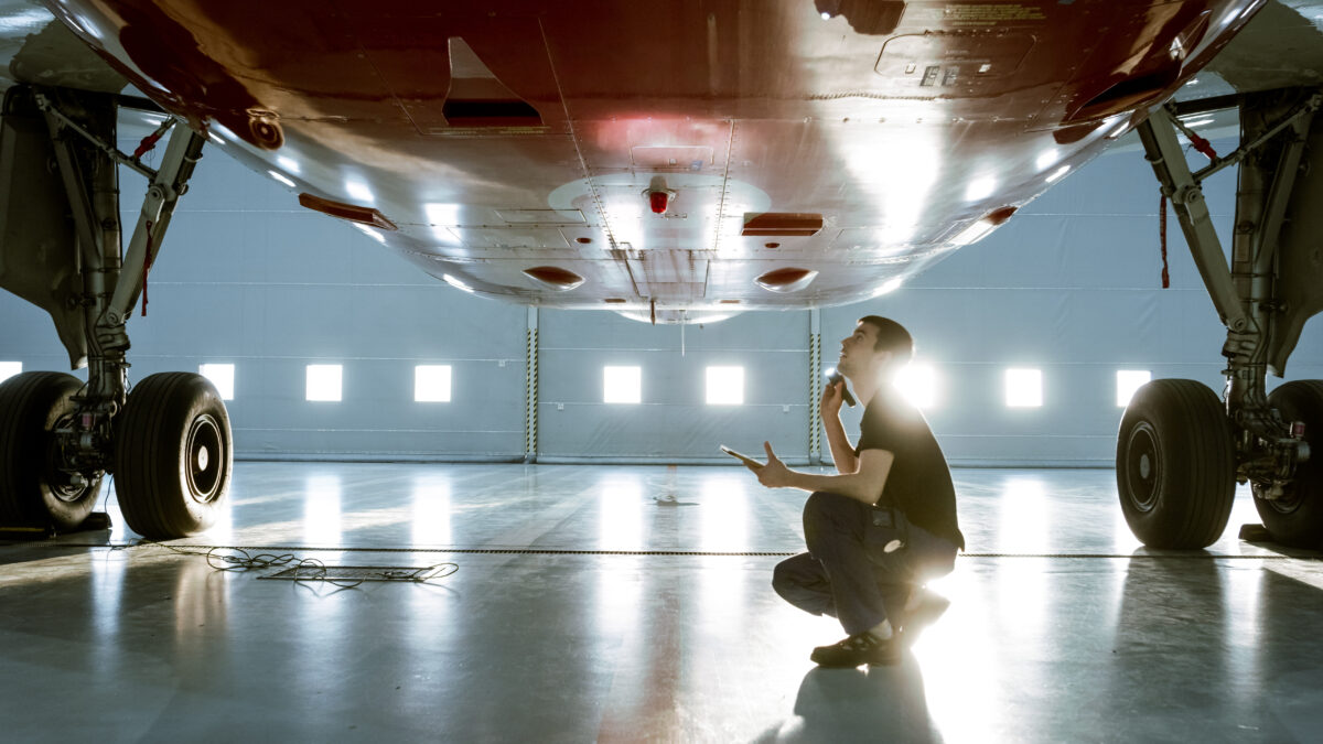 Aerospace technician inspecting the underside of an aircraft in a hangar, representing precision testing, safety checks, and performance validation, all core principles of JETS testing in aviation engineering.